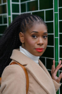 Safiya is standing in front of a green tiled wall with her head turned to the right to look directly down the camera lens. Safiya has long black hair, braided back into a curly ponytail. Safiya is wearing red lipstick, a white turtleneck and a beige coat. 