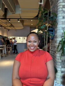 Sacha sits at a table, facing the camera and smiling. She is wearing a red shirt and has her dark hair tied back. 